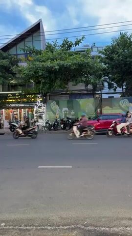 This media file captures a street scene in Đà Nẵng, Vietnam, likely during the late afternoon or early evening due to the soft, diffused light and overcast sky. The scene is bustling with activity.

The primary subjects are various vehicles and people navigating the street. A large white and red bus is prominently featured, moving from left to right across the frame. Several motorbikes are also visible, with people riding them, indicating common modes of transportation in the area. A red SUV is parked on the side of the road, and a white electric car with distinctive red and black racing stripes is driving past on the left.

The setting is an urban environment. On the right side of the street, there is a building with a modern, angled roof and large windows. Green trees line the sidewalk, and signs with yellow lettering are visible, suggesting commercial establishments like shops or restaurants. Some of these establishments are decorated with Christmas lights, hinting at a festive season. The road surface appears to be asphalt, and there are clear lane markings. The overall atmosphere is one of everyday urban life, with traffic and pedestrian activity.

The time of day is suggested by the twilight sky. The weather appears to be cloudy, contributing to the soft lighting. The location cues, such as the vehicle types (especially the electric car with Vietnamese branding) and the architectural style, point towards Vietnam. There is no overt display of strong emotions; the scene conveys a sense of normalcy and movement.

Visible text includes what appears to be a Vietnamese business name on the illuminated signs of the buildings on the right. The electric car also has branding on its side, including a "V" logo and the word "VINFAST," which is a Vietnamese automotive brand.