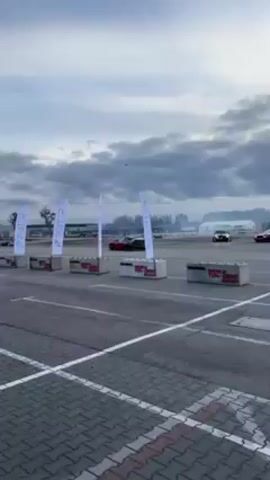 The video shows two cars drifting on an asphalt course marked by banners and concrete barriers. There are five white banners, each with a blue logo reading "PETER" vertically and additional text at the bottom. The concrete barriers beneath the banners also feature sponsor logos. The sky is overcast. A black and white car and a red car are drifting on the course, generating smoke. Other cars and people can be seen in the background. The camera is stationary.