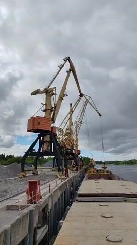 The video shows a dock scene with three large, stationary cranes loading cargo onto a ship. The cranes are beige with brown and black components. The cranes are positioned along a concrete dock, and a pile of gravel sits nearby. The ship is docked adjacent to the concrete dock and appears to be filled with some other type of material. The sky is overcast.