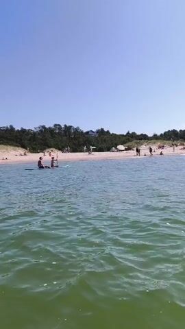 The video opens with a wide shot of a calm, green ocean under a bright blue sky. A sandy beach with scattered people and umbrellas stretches across the background. Several people are visible in the water, some on paddleboards. The camera then pans slightly to the right, showing more of the beach and the ocean. The perspective shifts to a first-person view, showing the front of a pink paddleboard with a foot visible. The paddleboard is moving through the water, creating small splashes. The beach and the sky remain visible in the background, with the sun glinting off the water's surface. The video concludes with a continued view of the paddleboard moving through the ocean towards the crowded beach.