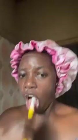 A person is seen from the shoulders up, facing the camera directly, brushing their teeth with a yellow-handled toothbrush. The person is wearing a pink patterned shower cap. Their face is covered in a white soapy substance. They have dark skin, dark eyelashes, and their eyes are squinted. The background includes light brown tiled walls and a dark colored doorway. The person vigorously moves the toothbrush across their teeth and tongue, making exaggerated facial expressions.