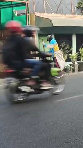The video shows a street sweeper in a blue and yellow shirt, dark pants, and a helmet, sweeping the side of a road with a broom. The sweeper is carrying a large sack over their shoulder. The scene appears to be in a tropical or subtropical urban environment, with lush green plants visible in the background. A motorcycle is seen passing by in the foreground, and another person wearing a white headscarf is partially visible in the frame. The buildings in the background suggest a mix of residential and commercial properties. The overall lighting indicates it is daytime, likely morning or late afternoon.