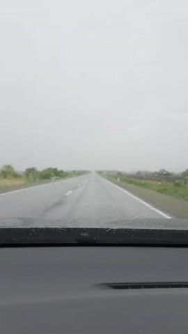 A dashcam view captures the perspective from inside a vehicle driving on a wet, two-lane asphalt road under an overcast sky. The road stretches straight ahead, marked with white lines, and is bordered by green vegetation, including bushes and distant fields, as well as white marker posts. The windshield wipers are audible, indicating active rain or recent precipitation, causing the road surface to reflect the dull light. The scene suggests a calm drive through a rural area in Бабстово, Russia, during daytime under cloudy weather conditions. No people or visible text are present.