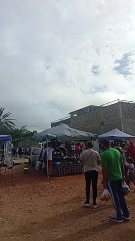 The video depicts an outdoor market scene with multiple stalls and people browsing. In the foreground, three individuals walk away from the camera. The person in the center, wearing a green shirt and dark pants, carries a plastic bag. To their left, a woman in a light-colored shirt and dark pants also carries a plastic bag. A man in a red shirt walks from right to left in the middle ground. Several stalls with tents are visible, displaying various items, likely clothing and other goods. People are gathered around these stalls. In the background, a large, unfinished concrete structure is visible, along with trees and a cloudy sky. The ground appears to be dirt or gravel.