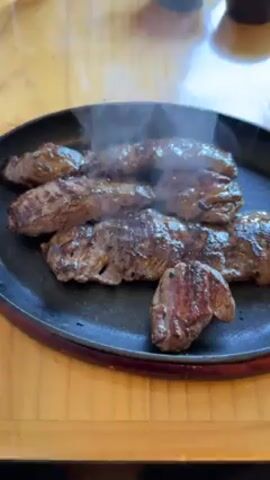 The video shows a black cast-iron plate filled with multiple pieces of cooked meat, probably steak. The meat is arranged on the plate, with steam rising from it, suggesting that it is freshly cooked and still hot. The plate is sitting on a wooden table, possibly in a restaurant setting. The lighting is natural and bright, highlighting the cooked texture and appearance of the meat. The camera appears to be handheld and the focus is on the plate of meat. There are some blurry objects in the background, but the main focus is the sizzling meat.