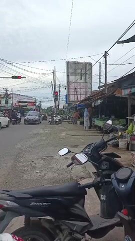The video shows a street scene with moderate traffic. In the foreground, a black scooter is parked. The street is paved with gravel and asphalt. Several cars and motorcycles are visible on the road, some in motion and others stopped at a traffic light. On the right side of the street, there are market stalls with awnings and people selling produce. The sky is overcast. Utility poles with numerous wires are visible throughout the scene.