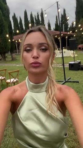 A blonde woman in a light green dress poses for a selfie in an outdoor grassy area. She is smiling and her hair is pulled back in a ponytail. In the background, there are lights strung across trees, chairs, and what appears to be a building. There are also speakers and stands visible. The woman moves her arm and spins around in a couple of frames.