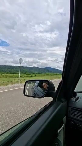 The video shows a view from inside a car, looking out the passenger side window. A side-view mirror reflects the interior of the car, showing a person's hand holding a phone, presumably taking the video. The car is driving on a paved road through a rural landscape with green fields and rolling hills in the background. The sky is partly cloudy. The video is shot in landscape orientation and has a timestamp at the beginning.