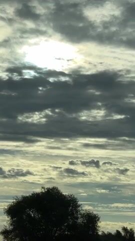 The video pans across a cloudy sky. The sun is visible behind a layer of clouds, casting a bright light. Below the sky, there is a silhouette of trees and vegetation. A road is visible in the foreground, with lane markings. There are also streetlights and traffic signals in the distance. The overall impression is of a vast, open landscape under a dramatic sky.