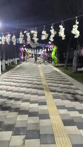 The video captures a park pathway at night, illuminated by overhead lights strung with white floral decorations and colorful balloons. The pathway is paved with gray and black blocks, with a yellow tactile paving strip running down the center. Several individuals, some accompanied by dogs, are seen walking in the distance. Additional decorations, including a person in a green and yellow costume, line the path. The surrounding environment includes trees with white-painted trunks and grassy areas.