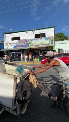A man wearing a white tank top, green pants, and a cap is pushing a three-wheeled cart laden with various items, including bags and what appear to be personal belongings. He is on a paved street with buildings lining the right side. One building has signage that reads "BALASTOS DE VENEZUELA" and "ELECTRIC ER." Cars are visible on the street, parked and in motion, and power lines crisscross overhead against a blue sky with scattered clouds. The man is pushing the cart away from the camera, moving down the street.