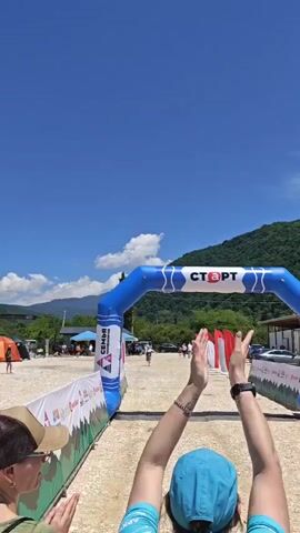 Two women stand in the foreground, clapping and cheering, facing away from the camera. In the background, a blue inflatable arch with the word "СТАРТ" in white letters spans a dirt path. A person in a white and black outfit runs towards the arch. Mountains and a blue sky are visible in the distance.