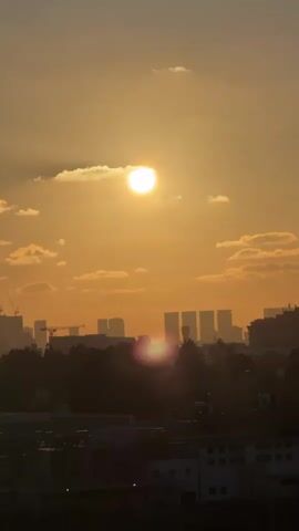 The video shows a time-lapse of the sun setting over a city. The sun is a bright orb in the sky, surrounded by clouds. Silhouettes of buildings and trees are visible in the foreground. The sky transitions from a bright yellow to a deep orange, indicating the progression of sunset. There are no people or animals visible in the video. The overall scene depicts a peaceful and serene urban landscape at dusk.
