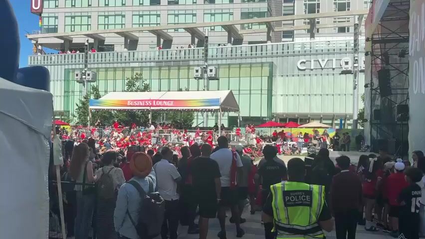 The video shows a crowd of people gathered outdoors during the daytime. In the background, there is a modern building with large windows and a sign that says "CIVIC". In front of the building, there is a temporary structure with a white tent and a banner that reads "BUSINESS LOUNGE" with a rainbow-colored flag garland. A stage is set up under this tent, with people performing or interacting in front of a seated audience behind a metal barrier. Many people in the crowd are wearing red and white clothing, suggesting a shared event or team affiliation. Some individuals are holding flags, including what appears to be a Canadian flag. There are also security personnel visible, including one wearing a high-visibility yellow vest with "POLICE" written on it. The overall atmosphere seems festive, with people engaged and observing the activities on stage.