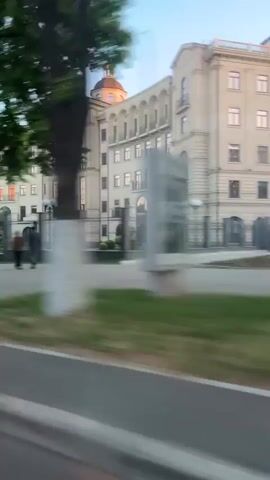 The video is shot from a moving vehicle and shows a multi-story building with a light facade, multiple windows, and a decorative roofline. The camera pans slightly to the right. A tall tree with green foliage partially obscures the view of the building. In front of the building is a black metal fence, a paved area, and a small guard house or kiosk. People are blurred in the background. The sky is light blue. The overall impression is of a sunny day in a formal or institutional setting.