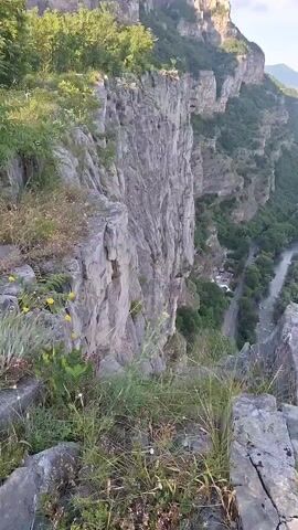 A drone flies over a dramatic cliffside overlooking a winding river and a road. The camera pans from a close-up of rocky terrain and wild vegetation to a panoramic view of the canyon. Lush green trees cling to the steep rock faces, and the river flows through the valley below. The sky is a pale blue with a few wispy clouds.
