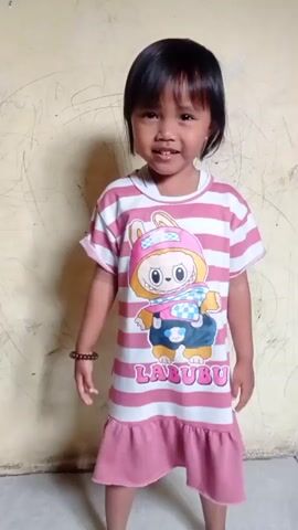 A young girl wearing a pink and white striped dress with a cartoon bunny is standing in front of a cream-colored wall with scuff marks. She turns to her right and walks towards an open doorway. Another child, on their hands and knees, is visible within the doorway. The first girl smiles and appears to be interacting with the second child.