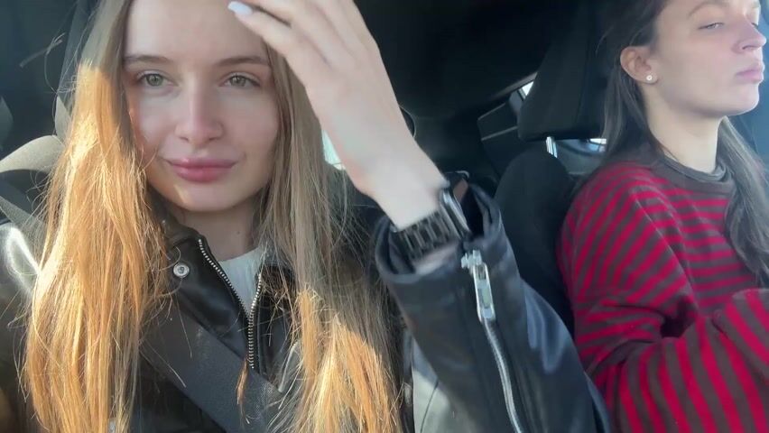 The video shows two young women in a car, presumably driving. The woman in the passenger seat has long blonde hair and is wearing a black leather jacket. She looks into the camera and briefly dances her fingers. The driver is wearing a red and black striped shirt and is focused on driving. The sun is shining brightly outside, indicating it's likely daytime. The interior of the car is modern, with black seats. The video appears to be shot from the passenger's perspective.