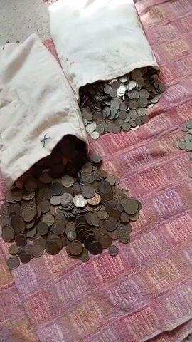 The video shows two cloth bags overflowing with coins. The coins are spread out on a pink and white striped fabric. Some coins have spilled out of the bags onto the fabric. The coins appear to be of various sizes and colors, suggesting they are of different denominations and possibly from different countries or eras. The lighting is natural, coming from above. The background is out of focus but appears to be a textured surface. The camera pans slowly across the scene, giving a close-up view of the coins.