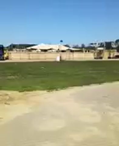 A wide shot shows a large, light-colored tent structure with a tall, thin pole extending upwards, surrounded by a low, light-colored fence. In front of the tent, there is a patch of green grass and a dirt path. To the left of the tent, a large, dark truck drives across the frame, kicking up dust. The sky is clear and blue.