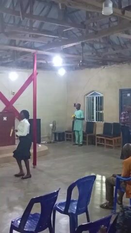 The video shows the interior of a church or similar gathering place. A woman in a white blouse and dark skirt is walking on a stage. A man in a light green traditional outfit is standing near a window, holding a yellow object. Several blue plastic chairs are in the foreground, and there are other chairs and a fan near the window. The room has a corrugated metal roof with exposed beams and several light fixtures. There's a cross-shaped decoration with red fabric on the stage, and what appears to be a speaker is also visible. The video seems to be taken from a low angle, looking up towards the stage and the man. There is music playing in the background.