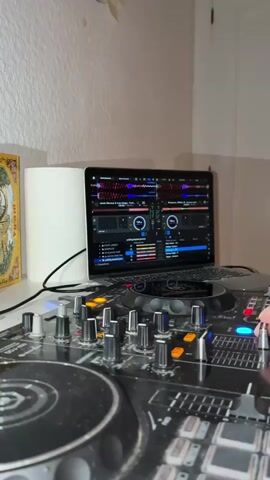 The video features a close-up shot of a DJ setup in an indoor environment. A black DJ controller is the primary focus, occupying most of the frame. The hand of a person with short, dark hair on their wrist is seen manipulating the knobs and buttons on the controller. In the background, a laptop displaying DJ software is visible. To the left of the laptop, a roll of white paper towels and a framed piece of art are placed. The wall behind the setup is painted white. The lighting in the room appears to be dim, emphasizing the lights on the DJ controller and laptop screen.