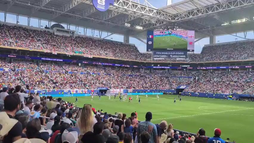 The video shows a crowded soccer stadium from the perspective of a spectator in the stands. The field is green, and there are players on the field, though they appear small due to the distance. A large screen at the far end of the stadium displays a close-up of the game, showing players in red and white uniforms. The stands are filled with a large, diverse crowd of spectators wearing various clothing and hats. The stadium has a modern roof structure with visible beams and lighting. The overall atmosphere appears to be one of an active sporting event with a significant audience.