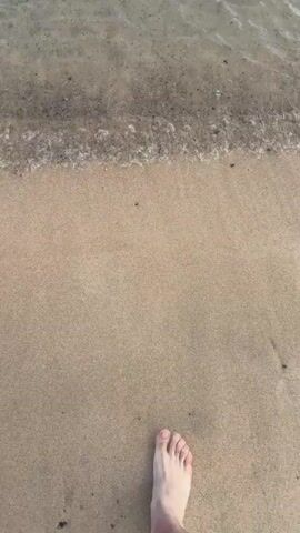 The video shows a single, light-skinned foot stepping from sand into clear shallow water. The foot enters the water creating a small splash. The water is clear enough to see the sandy bottom with some darker patches. The water ripples and moves away from the foot. The horizon line is visible with a clear blue sky above the water.