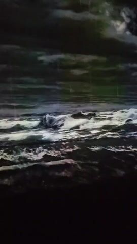 The video shows a projection of a stormy sea with dark, turbulent clouds in the sky. Rain is visible falling through the air. The waves are illuminated by a bright, possibly artificial light source, creating a dramatic and intense atmosphere. The scene is dark, with the exception of the illuminated waves and sky. There are no people or specific objects visible in the video, only the natural elements of a stormy sea and sky. The camera angle is slightly elevated, looking out over the water.