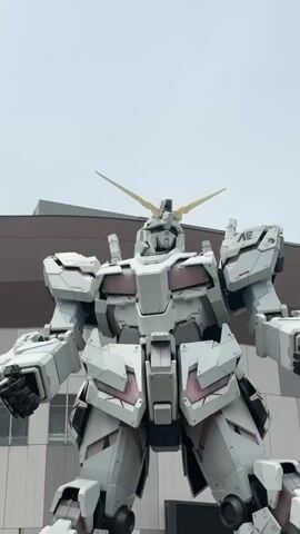 The video shows a low-angle shot of a massive white and gray robot standing outdoors. The robot is detailed, with panels, joints, and a prominent horn on its head. The background is a hazy sky with a building visible behind the robot's head and shoulders. The robot appears to be stationary.