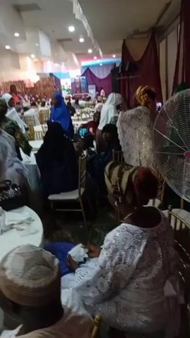 The video shows an indoor gathering with many people dressed in traditional attire. There are tables draped with white cloths, some with floral centerpieces and bottles of water. Several individuals are seated at the tables, while others are standing and interacting. The lighting is dim with pink and white accents. Some people are wearing headscarves and elaborate outfits. The overall atmosphere appears to be a formal event or celebration.
