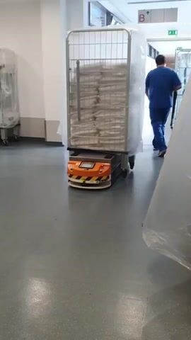 A robotic cart is moving down a hallway in what appears to be a hospital or healthcare facility. The cart is orange and black with a digital display and flashing orange lights. It is carrying a large metal cage filled with stacks of white linens, covered by a clear plastic sheet. In the background, a person wearing blue scrubs walks away from the camera. The hallway has a grey floor and white walls, with other metal carts visible in the distance.
