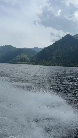 The video shows a view from the back of a moving boat on a large body of water. The water is dark and choppy with white foam trails from the boat's wake. In the distance, there are large, tree-covered mountains under a partly cloudy sky. The camera is angled downwards towards the water.
