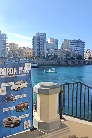 The video shows a scenic view of a harbor lined with buildings, a car rental sign in the foreground and several small boats in the water. The harbor water is a clear turquoise color. The camera pans right, revealing more of the harbor and the buildings lining it, a black metal railing in the foreground, and a set of steps leading down towards the harbor. The weather appears sunny and clear.