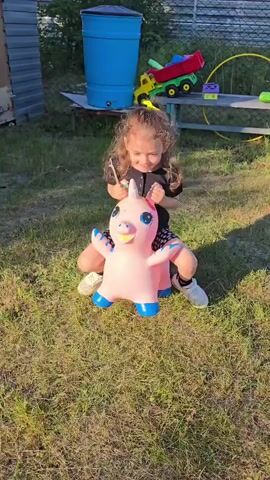A young girl with curly brown hair, adorned with yellow and red hair ties, sits astride a pink inflatable unicorn in a grassy outdoor area. She wears a black t-shirt and patterned shorts. Her hands grip the unicorn's ears as she bounces. The background shows a corrugated metal structure, a blue barrel, a toy truck, and a fence. The sun casts shadows on the ground.