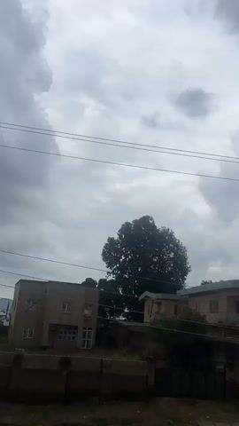 The video displays a cloudy sky above a residential area. Power lines crisscross the frame, supported by a utility pole in the foreground. Several buildings with varying architectural styles are visible, including a two-story house with white walls and multiple windows. Trees, including a large, leafy one, dot the landscape. In the middle ground, a street can be seen with a few people and motorcycles. The overall impression is of a somewhat overcast day in a densely populated urban or semi-urban environment.