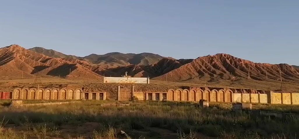 The video shows a wide shot of an outdoor scene with mountainous terrain in the background. In the foreground, there is a low-lying structure with a series of arched openings. Behind this, a longer, rectangular building is visible with a mural or painting on its facade. On top of this building, there is a statue or sculpture. The landscape appears arid with sparse vegetation. The lighting suggests it is either sunrise or sunset. There are no people or active events depicted in the video.