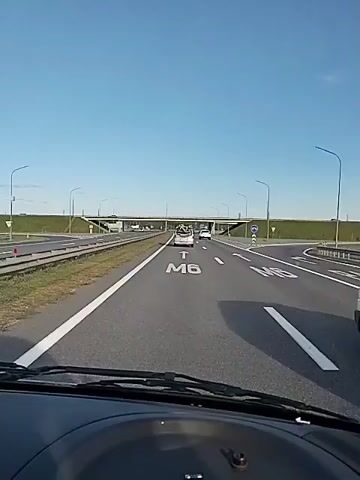 The video shows a first-person view from inside a vehicle traveling on a highway. The road is asphalt with white lane markings. In the foreground, the dashboard of the vehicle is visible. On the road ahead, large white letters "M6" are painted, indicating the highway number. There are other vehicles on the highway, including a white car ahead of the viewer's vehicle. In the distance, an overpass with streetlights crosses the highway. The sky is clear and blue. The overall environment appears to be a rural or suburban highway setting on a clear day. The audio consists of speech, the content of which is not directly related to the visual scene.