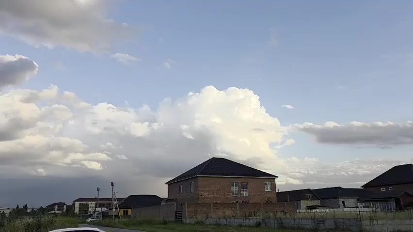 The video pans across a sky filled with white and gray clouds under a blue sky. In the foreground, there are some buildings and fences, including a brick house with a dark roof. There is also some greenery and a white car partially visible. In the distance, there are more buildings and what appears to be a mosque with a golden dome and a minaret. The scene suggests an outdoor, possibly suburban or rural environment.