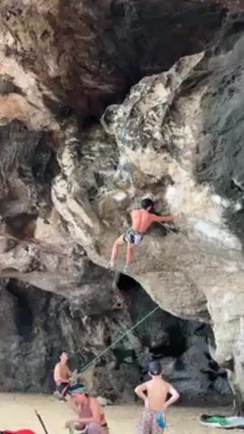 The video shows a shirtless rock climber ascending a rock face at a beach. He is wearing shorts and climbing shoes and is secured by a rope. The rock face is gray and white with interesting patterns. The background shows a sandy beach with several people. One person sits on the sand with climbing gear. Another man stands shirtless, while a child with a hat stands with their arms crossed. The sky is not visible, but the light suggests it is a sunny day. The scene conveys the activity of rock climbing in a natural beach setting.