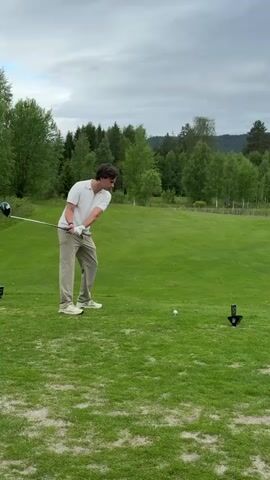 A man wearing a white shirt and tan pants swings a golf club at a golf ball on a green golf course. The sky is overcast and there are trees and a hill in the background. After the swing, the man watches the ball travel across the course. The camera pulls back to show the rest of the course and the surroundings.