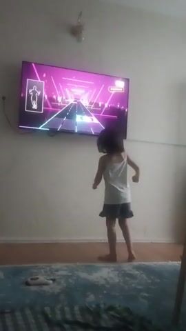 A young girl with dark hair, wearing a white tank top and a blue skirt, is standing on a blue rug in front of a large television screen mounted on a white wall. The television displays a video game with a pink and purple neon tunnel with a race track in the center, and a silhouette of a dancing figure in the upper left corner. The girl is facing the television and appears to be playing the game, as her body is positioned as if she is mimicking the dancing figure on the screen. She is barefoot. A white object, possibly a shoe, is on the rug near her feet. The room is simply furnished, with the focus being on the girl and the television.
