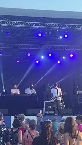 A live music performance is taking place on an outdoor stage. Three men are visible on stage. Two men are standing behind a DJ booth on the left side of the stage. One of them, wearing a white t-shirt, is operating the DJ equipment. The other man, also in a white t-shirt, is holding a microphone and appears to be singing or speaking into it. On the right side of the stage, a man wearing a white shirt and a baseball cap is playing an electric guitar. He is facing the audience and is in a dynamic pose. The stage is lit with blue spotlights and beams of light. In front of the stage, a crowd of people are watching the performance. The audience members are diverse in age and gender, and most are facing the stage. The overall environment suggests a summer concert or festival.
