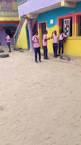 The video shows a sandy courtyard with a colorful, multi-story building in the background. Several young males, all wearing pink shirts and dark pants, are present in the courtyard and the building's ground floor entrance. Some are standing in small groups, possibly conversing. One individual is near the building's stairs, and another is closer to the camera, near a pile of tires. The building itself is painted in bright, contrasting colors, including yellow, blue, pink, and purple. The ground is covered in sand, with some tire remnants visible scattered around the courtyard. The overall scene appears to be an outdoor area of a school or similar institution.
