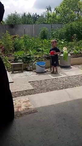 A woman in a black polka dot dress stands in a garden path facing away from the camera, her left hand on her hip. A young boy wearing a dark blue t-shirt and shorts stands to her right, holding a red watering can and facing the woman. He is positioned near a raised garden bed filled with plants. In front of the boy, to his left, is a blue and white bucket. Behind the boy and to his right is a tall, silver container. The garden path is made of grey concrete slabs and gravel, with a brick border on the left. The background features lush green plants, a metal fence, and trees. The sky is cloudy.