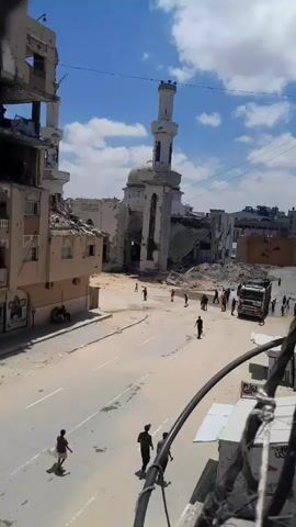The video shows a street scene in a damaged urban environment. In the background, a mosque with a tall minaret is visible, but its dome and sections of the building appear to be destroyed. Adjacent to the mosque, other buildings are also in ruins, with piles of rubble and debris. Several people are walking in the street and near the damaged structures. A large white truck with a black tank on the back is parked in the street. Some individuals appear to be carrying buckets or containers, and one person is interacting with the truck. The ground is dusty and appears to be paved. The sky is blue with some clouds, suggesting it is daytime.