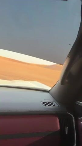 From the interior of a car, a desert landscape is visible through the windshield. Sand dunes stretch into the distance, with tire tracks marking the sand. Another vehicle is seen driving on the dunes, and the dashboard and a red seat are visible in the foreground.
