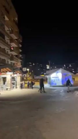 The video shows a night scene in a snowy town. There are people walking on a snow-covered path. On the left, there's a multi-story building with lights. To the right, there are small structures, some illuminated with blue and red lights. In the distance, lights from other buildings dot the dark sky. The camera seems to be moving slowly along the path.