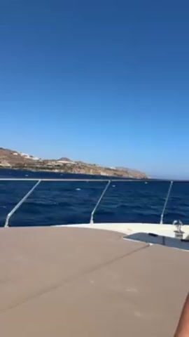 Here's a concise description of the video:

The video begins with a view from a boat, showing a coastline in the distance under a clear, blue sky. The camera then focuses on a woman who is lying face-down on the deck of the boat. She is wearing a swimsuit, sunglasses, and a pearl necklace. The boat is moving, as evidenced by the waves.