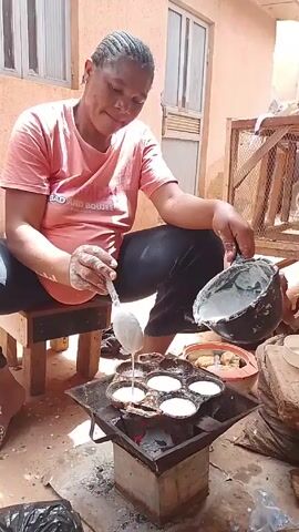 A woman is sitting on a wooden stool outside, cooking. She has dark hair styled in braids and is wearing a pink t-shirt and dark pants. She is pouring a white liquid from a black pot with her right hand, using a spoon in her left to assist. The liquid is going into a flat, circular pan with multiple indentations, which is positioned over a small charcoal stove. The stove is a square metal frame with a concrete base, and embers are visible inside. The background consists of a building with windows, various objects like a caged structure, a container with more of the cooked food, and general outdoor detritus. The scene is brightly lit with natural daylight.